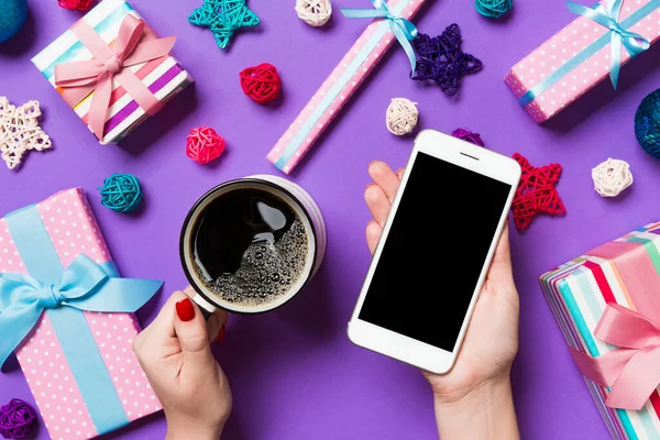 Bir elinde telefon, diğer elinde bir fincan kahve olan bir kadının mor arka planda görüntüsü. Noel süslemeleri ve oyuncakları. Yeni yıl tatili konsepti. Model.