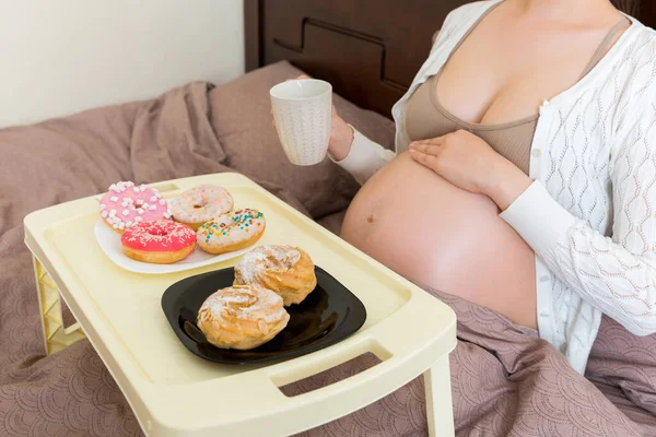 Hamile kadının yakın çekimi evde yatakta kahvaltı yapmaktır. Çörek ve kek tepsisi. Hamilelik sırasında hamur işi yemek..