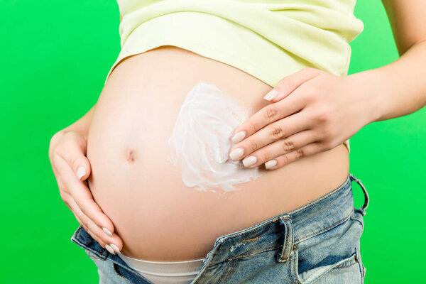 Close up of pregnant woman in unzipped jeans applying moisturizing cream on her belly against stretch marks at colorful background with copy space. Skin care concept.