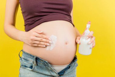 Açık kotlu hamile kadının, renkli arka planda fotokopi aletiyle oluşan çatlaklardan kaçınmak için göbeğine nemlendirici losyon sürerken yakın plan. Hamilelik konsepti sırasında cilt bakımı.