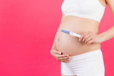 Beyaz iç çamaşırlı hamile bir kadının, pembe arka planda iki şeritli pozitif gebelik testi yaparken görüntüsü. Sağlıklı hamilelik. Boşluğu kopyala.