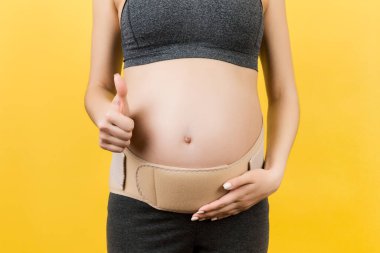 Hamile bir kadının baş parmağını göstermesi ve elastik hamilelik bandajı takıp sarı arka plandaki ağrının geçmesini sağlamak için fotokopi çekmesi. Ortopedik abdominal destek kemeri kavramı.