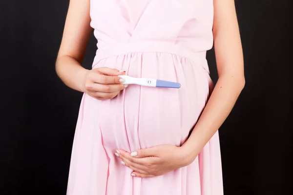 Close up of positive pregnancy test against pregnant woman\'s belly at black background. Future mother in white underwear. Baby expecting. Copy space.