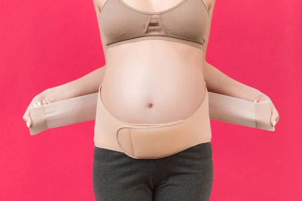 Close up of pregnant woman dressing elastic maternity band at pink background with copy space. Orthopedic abdominal support belt concept.