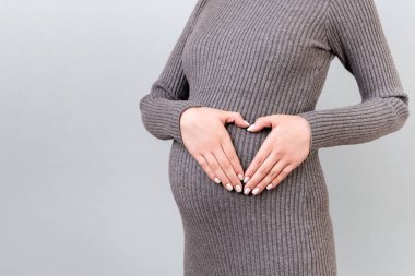 Gri arka planda bebek çarpması sonucu elini kalp şeklinde tutan hamile bir kadın görüntüsü. Annelik kavramı. Boşluğu kopyala.