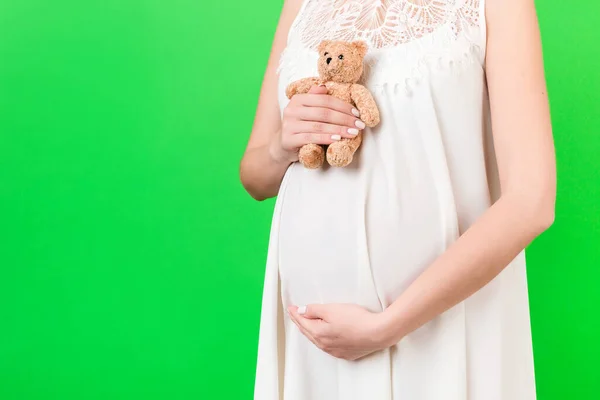 緑色の背景でお腹にテディベアを背負ったまま白いドレスを着た妊婦のクローズアップ 若い母親は赤ん坊を期待している スペースのコピー — ストック写真