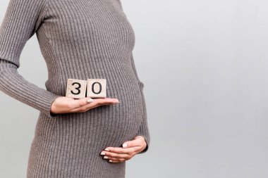 Hamile bir kadının ellerinde 30 haftalık hamile bir kadının gri arka planda gri bir elbise giydiği küp görüntüsü. Teslimat geliyor. Boşluğu kopyala.