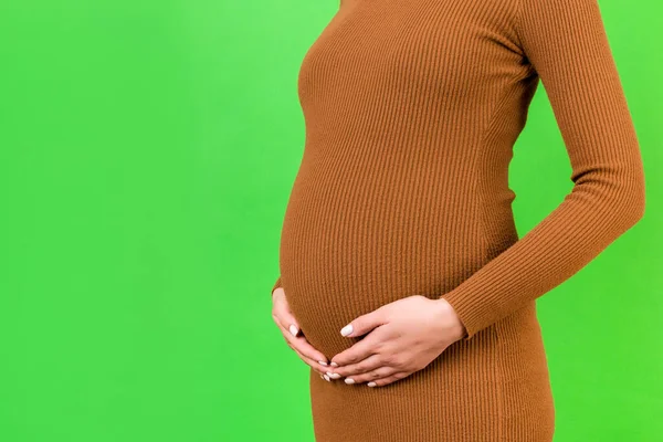 怀孕妇女抱着她的腹部的剪影 母亲穿着绿色背景的褐色连衣裙 未来的父母正在等她的孩子 复制空间 — 图库照片