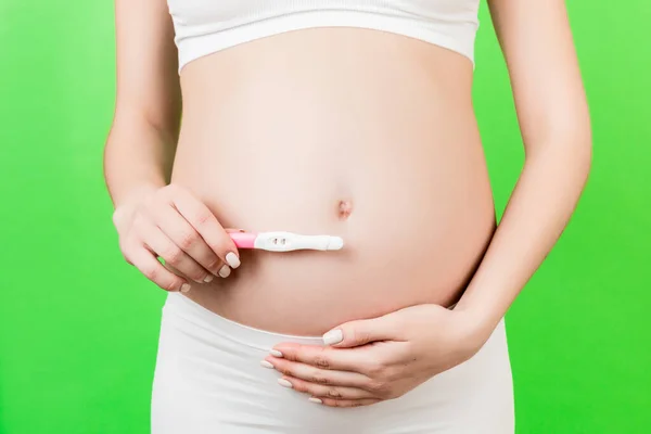 Close up of positive pregnancy test against pregnant woman\'s belly at green background. Future mother in white underwear. Baby expecting. Copy space.