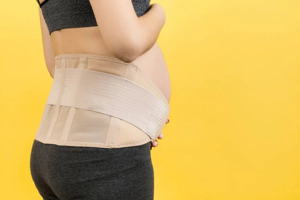 Back view of pregnant woman wearing pregnancy belt at yellow background with copy space. Close up of orthopedic abdominal support belt concept.