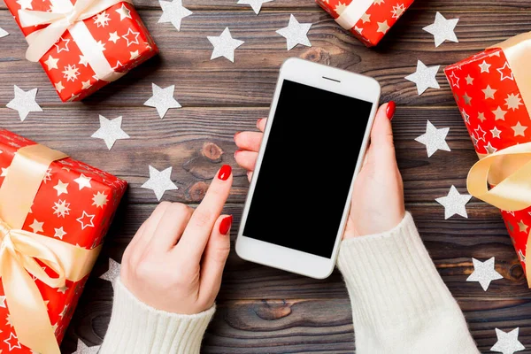 Top view of a woman holding a phone in her hand on wooden New Year background made of gift boxes and snowflakes. Christmas holiday concept. Mockup.