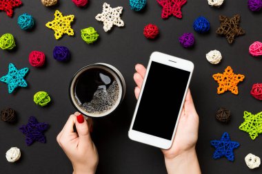 Bir elinde telefon, diğer elinde kahve olan bir kadının siyah arka planda görüntüsü. Noel süslemeleri ve oyuncakları. Yeni yıl tatili konsepti. Model.