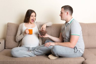 Mutlu koca, hamile karısını çikolatalı gevrek ve meyve suyuyla besliyor. Evdeki kanepede dinleniyor. Müstakbel baba, anneyi bekliyor..