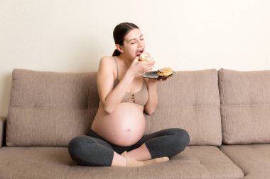 Genç hamile kadın evdeki kanepede dinlenerek lezzetli pastalar yemekten hoşlanıyor. Hamilelik sırasında sağlıksız beslenme.