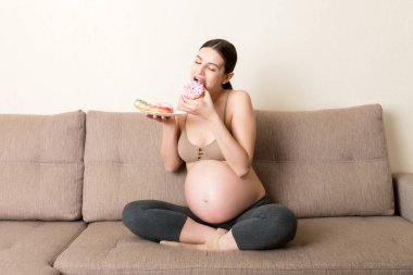Hamile kadın kanepede dinlenirken bir sürü çörek yiyor. Hamilelik sırasında sağlıksız diyet.