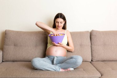 Kanepede oturan hamile kadın tuz arzusu yüzünden cips yiyor. Hamilelik sırasında sağlıksız abur cubur.