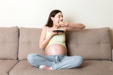 Aç hamile kadın evdeki kanepede büyük bir zevkle hamur işi yiyor. Mutlu hamilelik kavramı.