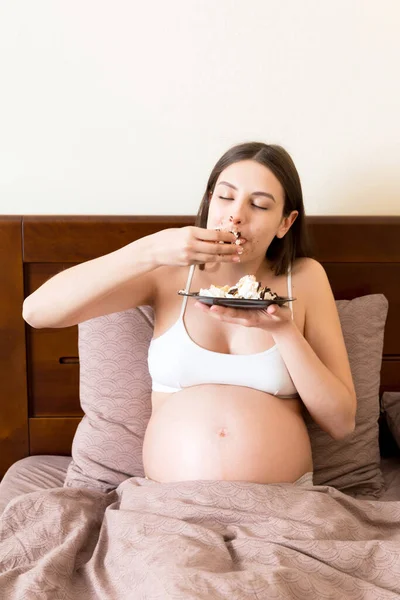 Hungry pregnant woman relaxing in bed is eating greedily a piece of cake and has a dirty mouth. Expecting mother can\'t stop eating. Strong appetite during pregnancy concept.