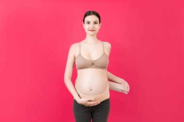 Pembe arka planda ağrıyı azaltmak için ortopedik bandaj giyen hamile kadının portresi. Ortopedik abdominal destek kemeri kavramı.