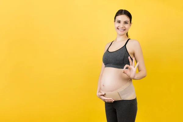 Portrait of pregnant woman wearing pregnancy bandage on her belly to make the backache go away and showing okay gesture at yellow background. Copy space. Orthopedic abdominal support belt concept.
