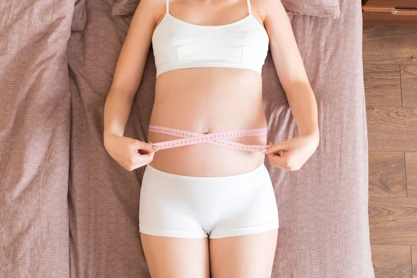 Young beautiful pregnant girl measuring her belly with a tape in home on bed. Healthy pregnancy concept.