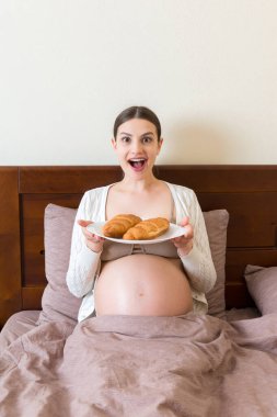 Yatakta dinlenen hamile bir kadın evde bir tabaktan kruvasan yiyor. Sağlıksız diyet. Hamilelik konsepti sırasında kilonu kontrol et.