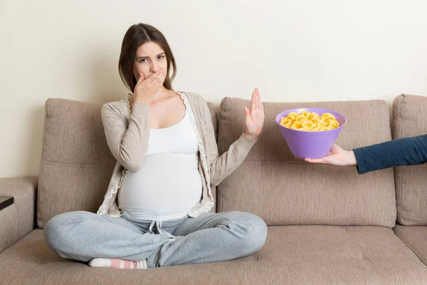 Pregnant woman sitting on the sofa refuses to eat unhealthy snacks. Stop to the junk food during pregnancy concept.