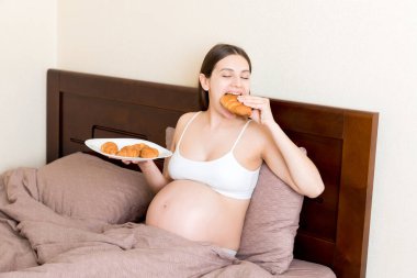 Yatakta dinlenen hamile bir kadın evde bir tabaktan kruvasan yiyor. Sağlıksız diyet. Hamilelik konsepti sırasında kilonu kontrol et.