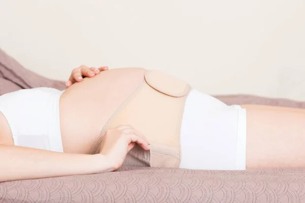 特写镜头孕妇在家里的床上用绷带包扎肚子 整形外科腹部支持带 — 图库照片