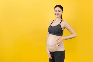 Sarı arka plandaki sırt ağrısını fotokopi aletiyle azaltmak için hamile kadınların giydiği elastik doğum bandı portresi. Ortopedik abdominal destek kemeri kavramı.