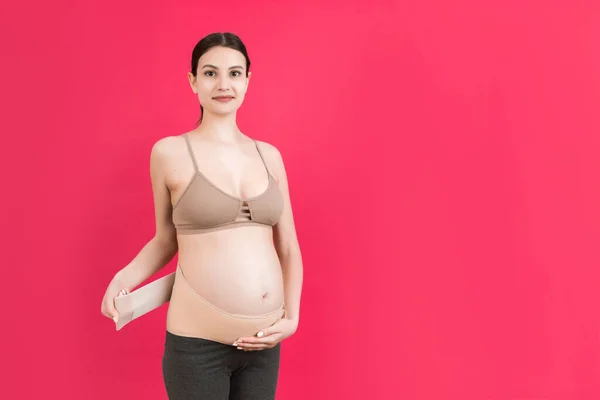 Portrait of pregnant woman dressing pregnancy corset on the third trimester at pink background with copy space. Orthopedic abdominal support belt concept.