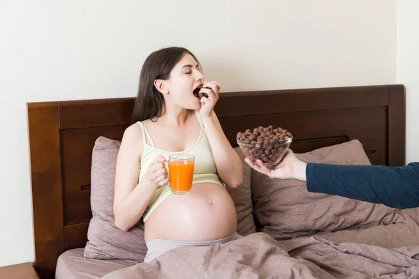 ベッドでリラックスした妊婦はチョコレートシリアルボールを食べ ジュースを飲んでいます 夫は妻に食事を与えている ドライブレックファーストチョコがポップ 妊娠中の朝の飢餓の概念 — ストック写真