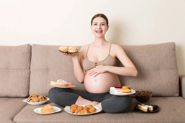 Hungry pregnant woman sitting on the sofa is eating a lot of unhealthy food such as donuts, cakes, chocolate cereal balls, croissants and cookies. Sweet cravings during pregnancy concept.