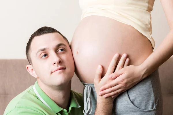 若いひげを生やした男彼の耳を彼の妊娠中の妻腹部に近づけて それを聞く — ストック写真
