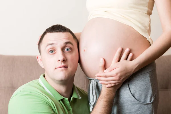 一个留着胡子的年轻人把耳朵贴在他怀孕的妻子的腹部 图库照片