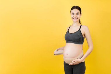 Hamile kadın portresi elastik annelik bandını sarı arka planda fotokopi aletiyle giyiyordu. Ortopedik abdominal destek kemeri kavramı.
