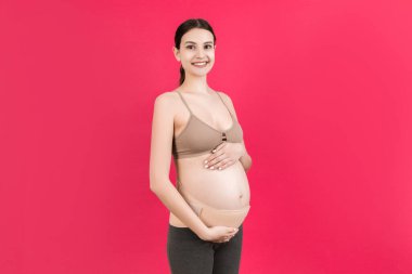 Portrait of pregnant woman wearing pregnancy corset against backpain at pink background with copy space. Orthopedic abdominal support belt concept. clipart