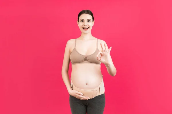 Portrait of pregnant woman wearing pregnancy bandage on her belly to make the backache go away and showing okay gesture at pink background. Copy space. Orthopedic abdominal support belt concept.