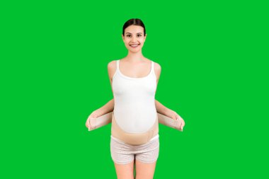Yeşil arka plandaki sırt ağrısını fotokopi aletiyle azaltmak için destekleyici bandaj takan hamile kadının portresi. Ortopedik abdominal destek kemeri kavramı.