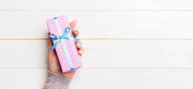 Kadının elleri çizgili hediye kutusunu tutuyor. Beyaz rustik ahşap arka planda renkli kurdele var. Noel konsepti ya da diğer tatil el yapımı hediye kutusu, kopya alanı olan konsept üst görünüm.