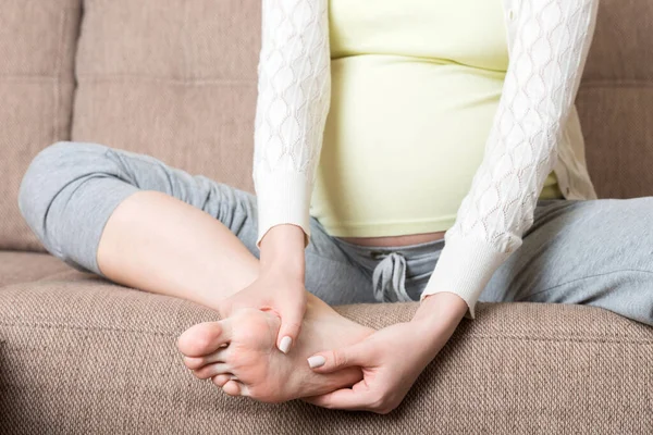 Leg cramps during pregnancy. Closeup of hands massaging swollen foot while sitting on sofa. — Stock Image Leg Cramps Pregnancy Closeup Hands Massaging Swollen Foot While Sitting Stock Image