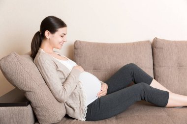 Müstakbel tatlı beyaz annenin oturma odasında kanepede uzanmış ve karnının üstünde el ele tutuşurken fotoğrafı..