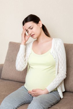 Kanepede oturan, başı ağrıyan hamile genç bir kadın. Hamilelik beklentisi kavramı.