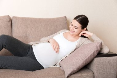Müstakbel tatlı beyaz annenin oturma odasında kanepede uzanmış ve karnının üstünde el ele tutuşurken fotoğrafı..