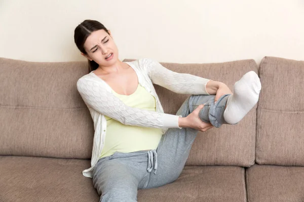 Leg cramps during pregnancy. Closeup of hands massaging swollen foot while sitting on sofa. — Stock Image Leg Cramps Pregnancy Closeup Hands Massaging Swollen Foot While Sitting Stock Image