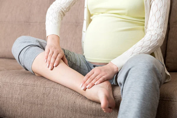 Leg cramps during pregnancy. Closeup of hands massaging swollen foot while sitting on sofa.