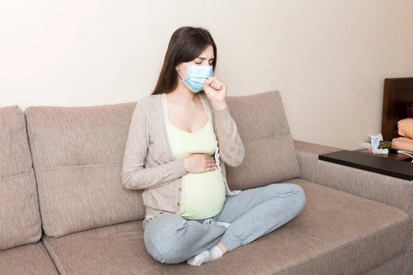 Pregnant woman in protection mask. COVID-19 Coronavirus is sitting on sofa. Prevent infection to the fetus.