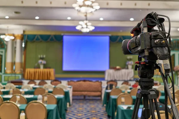 Siyah video kamera tripod üzerinde yakından var bulanıklık mavi ekran Projektör masa ve sandalye konferans salonunda sahnede arka plan olarak. İş ve seminer kavram başarılı.