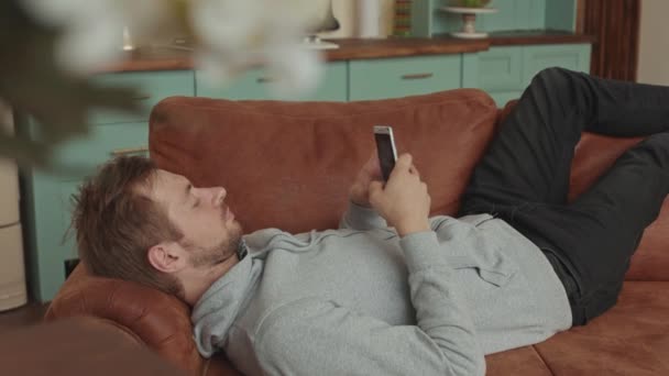 En man ligger på en soffa och använder en smartphone. Mysigt vardagsrum — Stockvideo