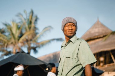 Rasta şapkalı bir adam arkasına bakıyor. Balili adam palmiye ağaçlarının arka planına gider.